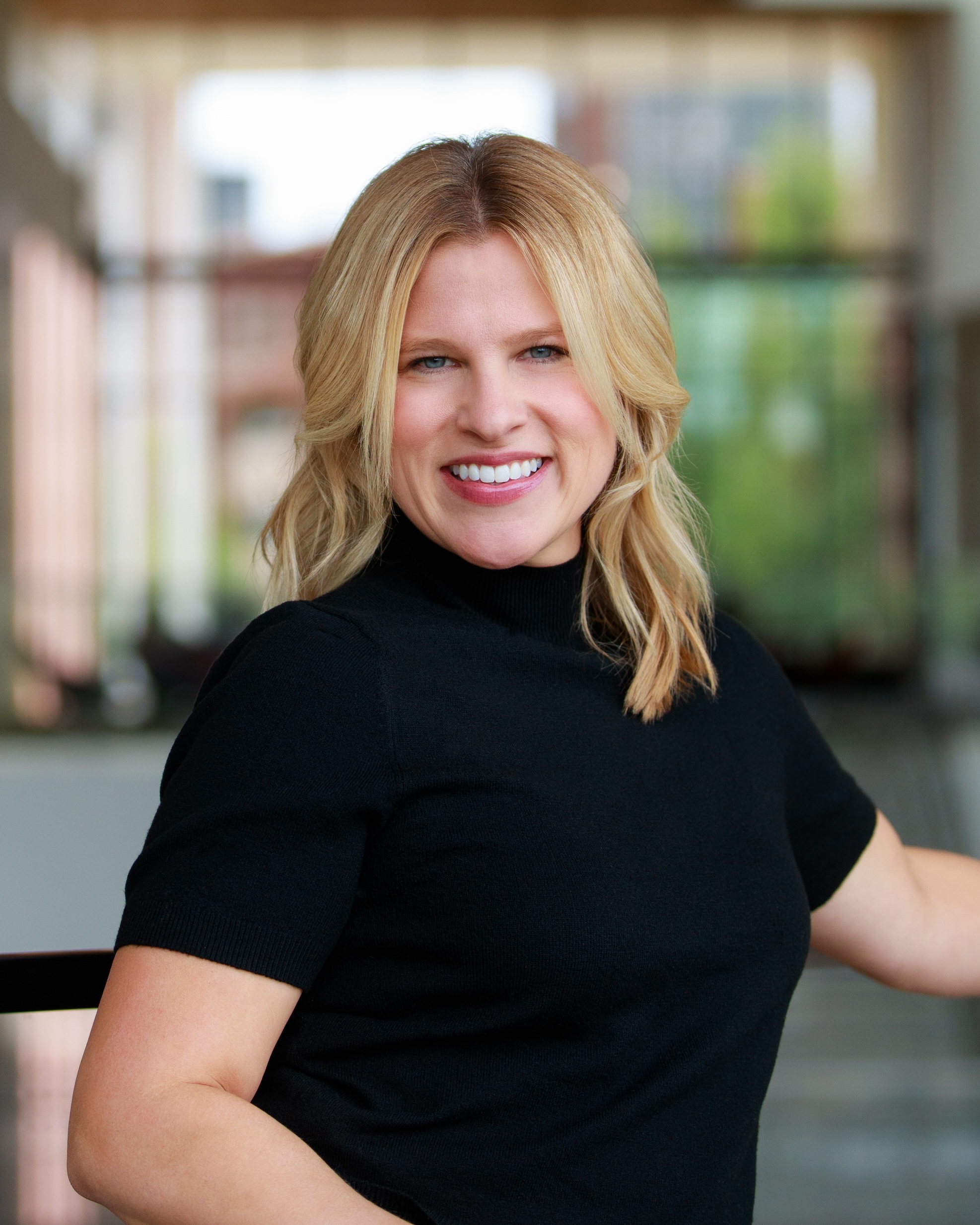 Portrait of Laura Winter indoors, wearing a short sleeve black top, photographed in front of large windows with a background showing interior architecture and greenery